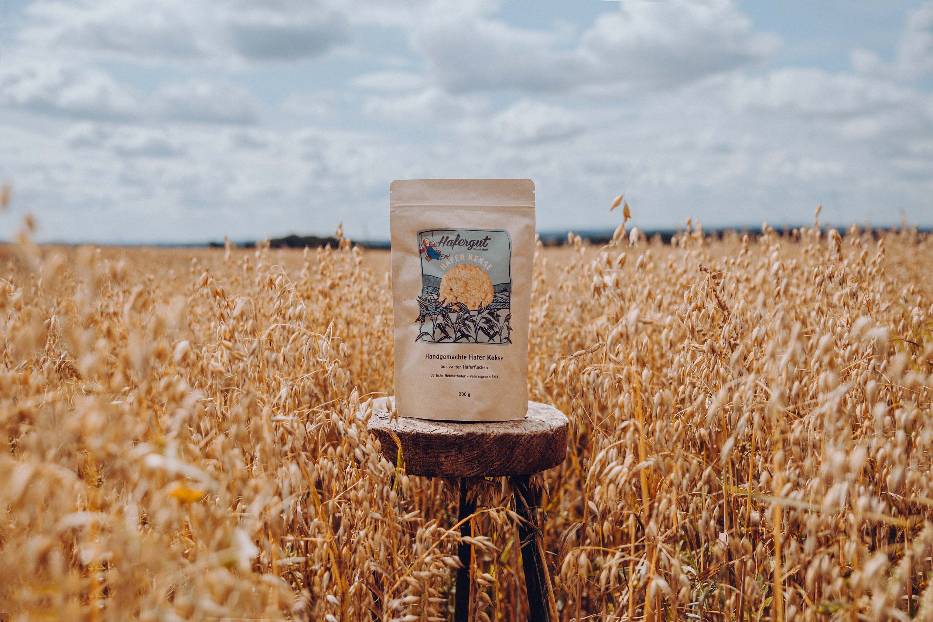 Eine Kekstüte steht auf einem alten Hocker aus Holz. Das Bild ist an einem bewölkten Tag, in Mitte eines Haferfeld aufgenommen worden.