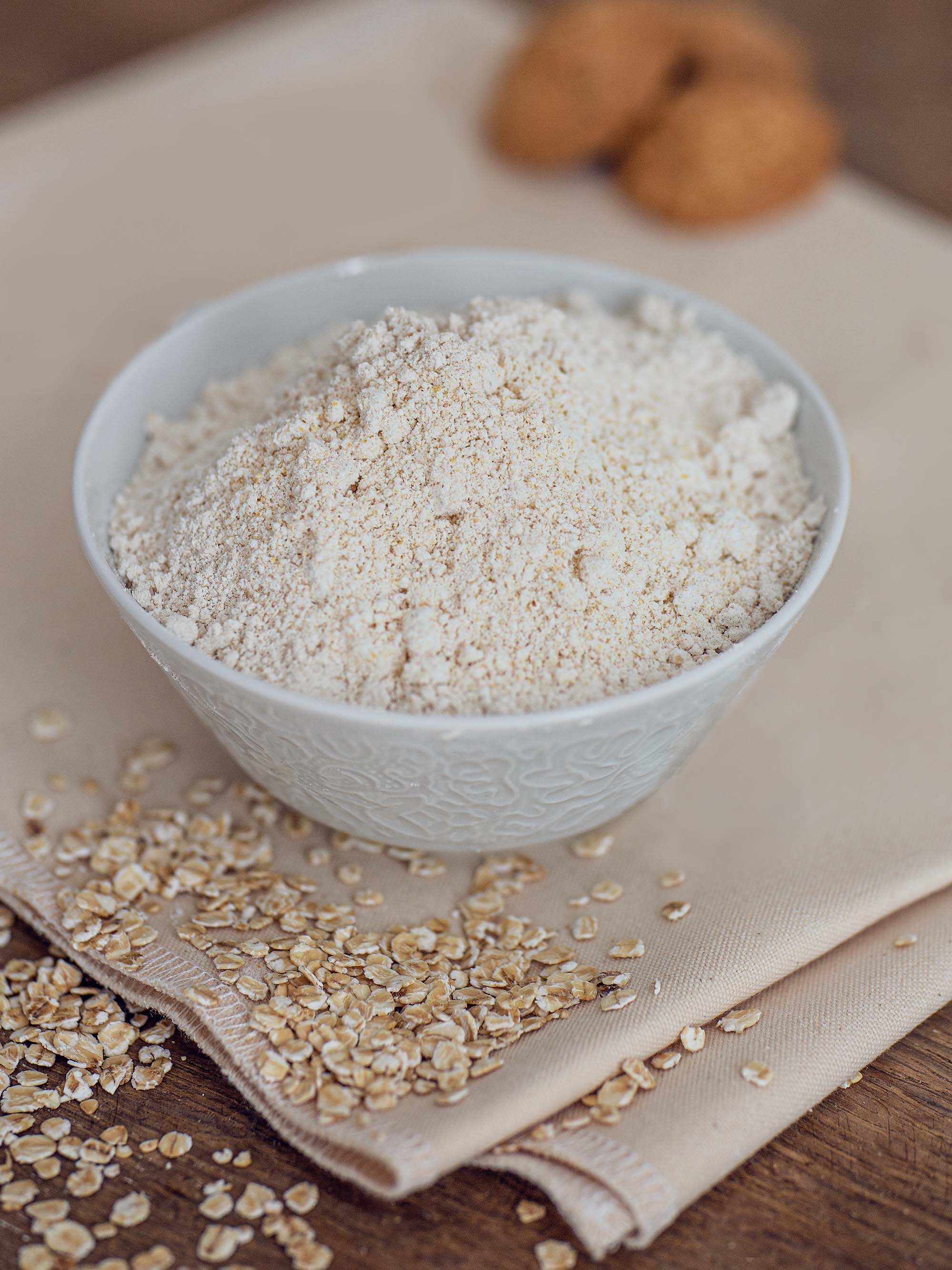 Hafervollkornmehl ist in einer Schüssel aufgetürmt. Man kann die Struktur des Mehls sehen und erkennt, dass es sich dabei um ein Vollkornmehl handelt. Unterstützend liegen Haferflocken um die Schüssel herum.