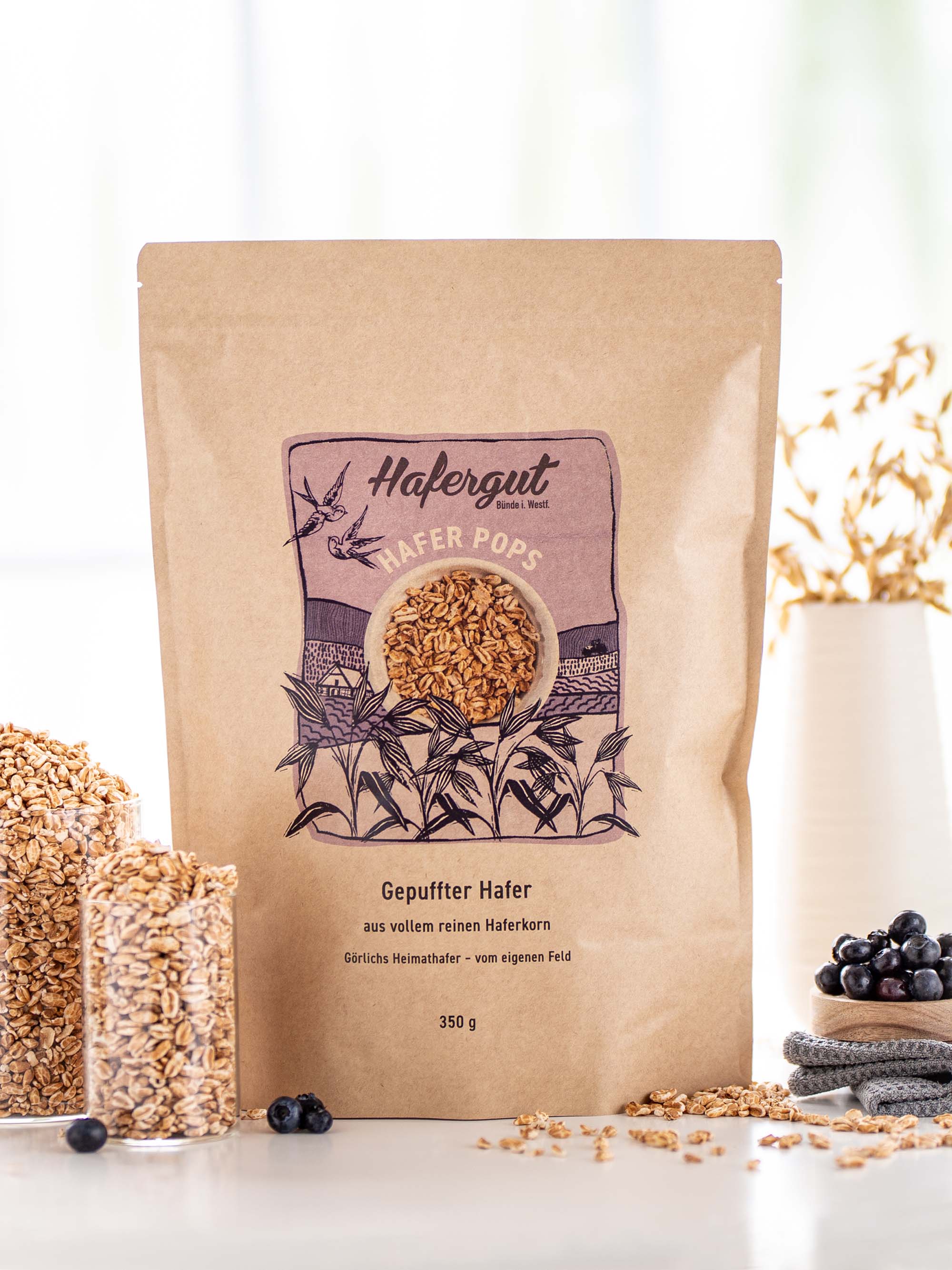 Haferpops sind in seiner Verpackung neben farblich passenden Blaubeeren, welche auf einem Holz-Schälchen drapiert sind, fotografiert. Links sind in zwei Glasgefäßen Hafer Pops präsentiert.