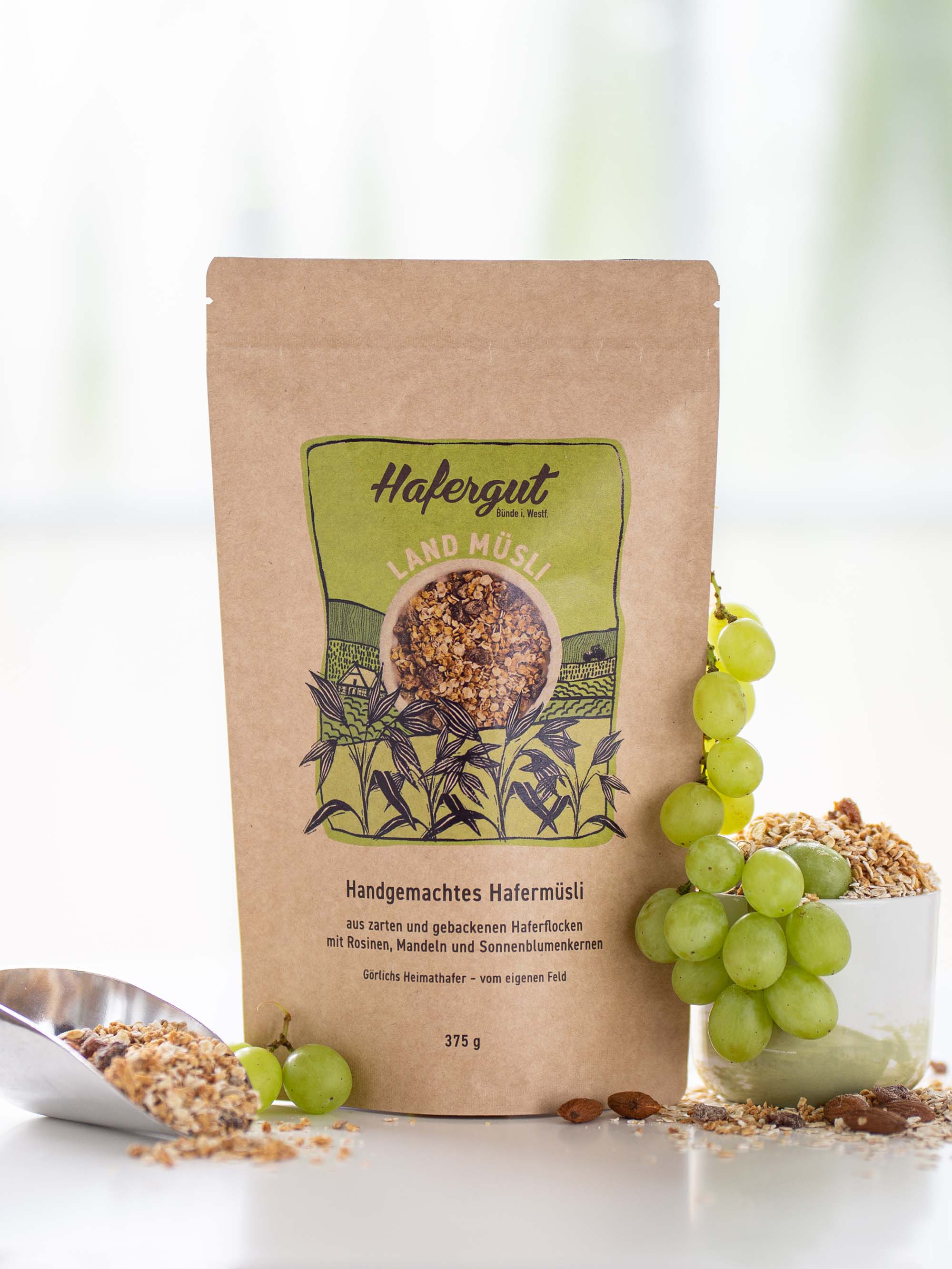 Das Produkt Land-Müsli mit Rosinen ist in seiner Verpackung als Produktbild fotografiert. Umrandet wird das Bild mit grünen, saftigen Weintrauben und verstreuten Landmüsli.