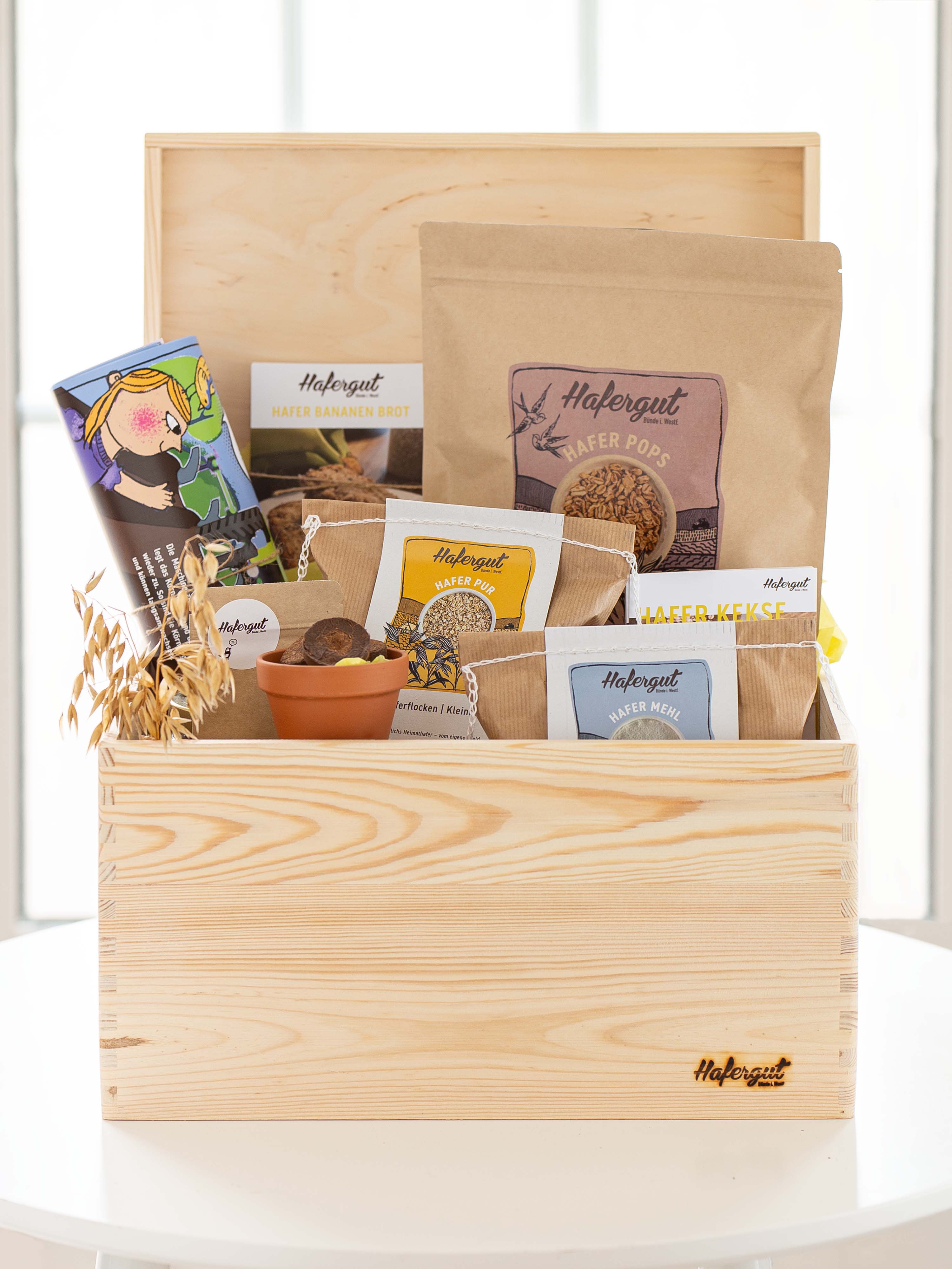 Zu sehen ist eine prall gefüllte Holzkiste mit vielen tollen Dinge für Kinder. Beinhaltet sind kindliche Poster, Rezeptkarten, Haferrispen, Anzuchttopf, Hafer Mehl, Hafer Flocken und Hafer Pops. Auf der hellen Holzkiste befindet sich unten rechts ein eingebranntes Hafergut Logo.