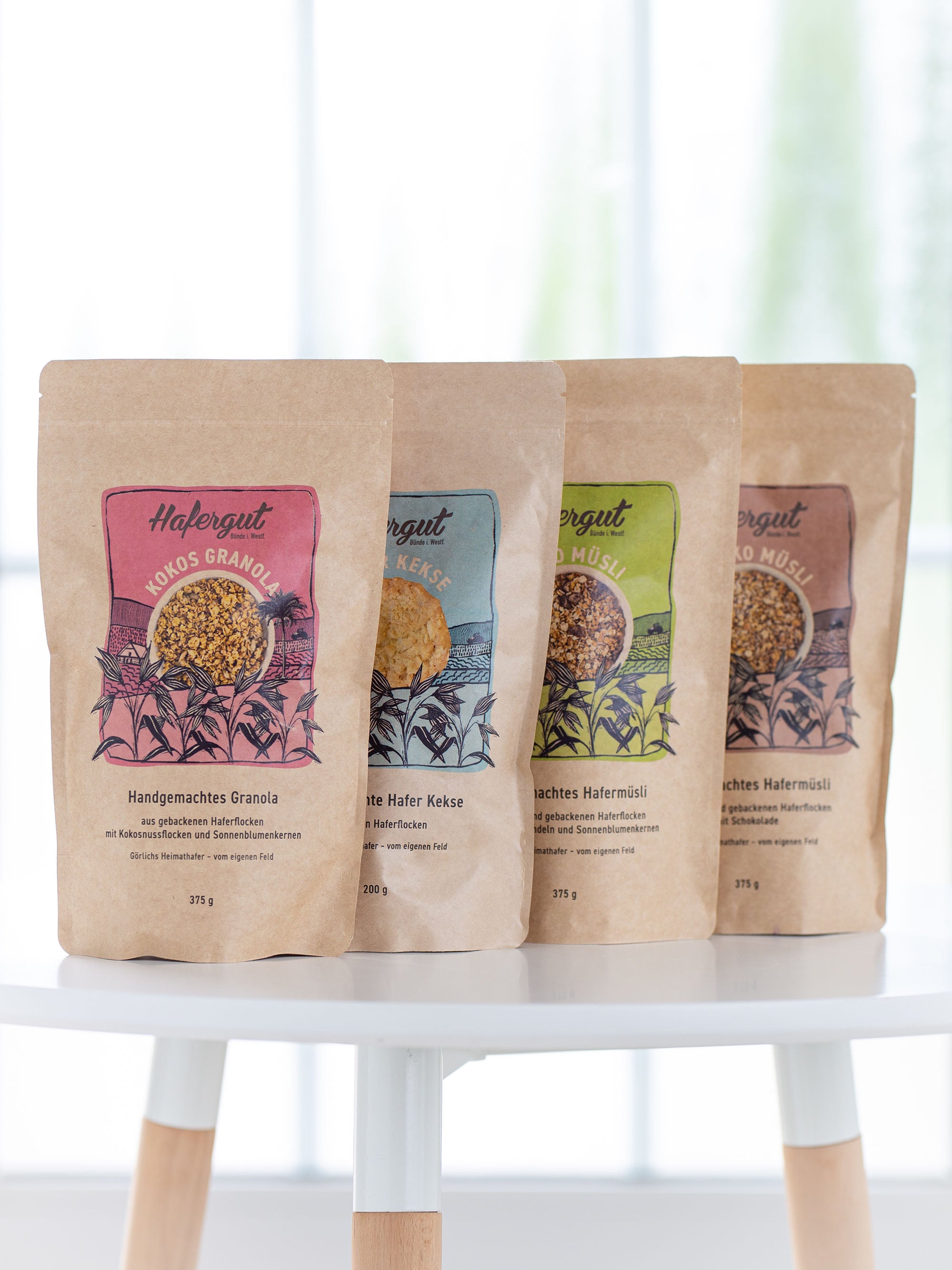 Auf einem Tisch stehen 4 verschiedene Tüten der Sorte Granola mit Kokos, Hafer Kekse, Land Müsli mit Rosinen und Schoko Müsli.