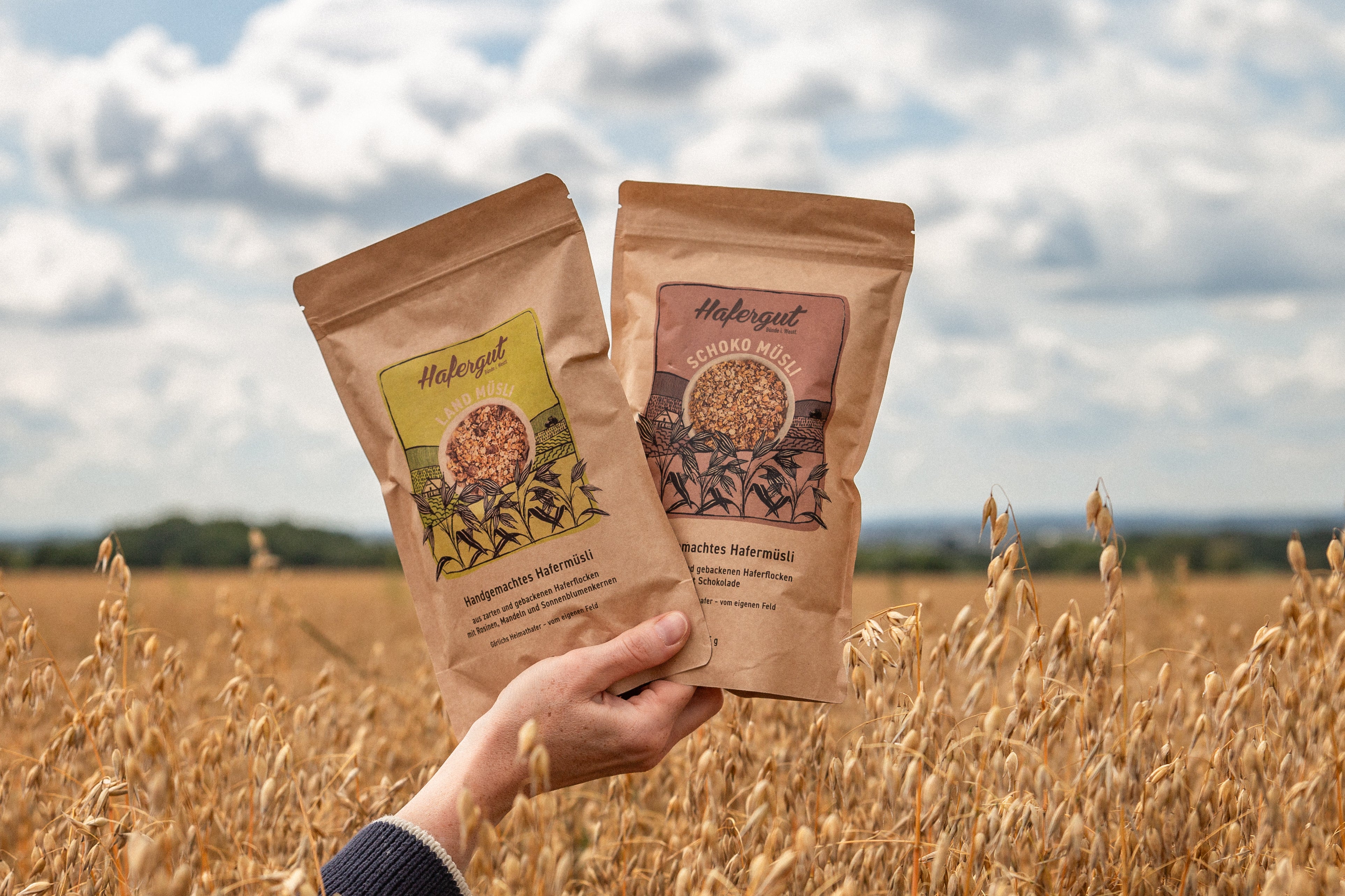 Eine Hand hält zwei verschiedene Müsli Tüten in der Hand. Sie präsentiert das Landmüsli und Schokomüsli. Das Bild ist an einem bewölkten Tag, in einem Haferfeld aufgenommen worden.
