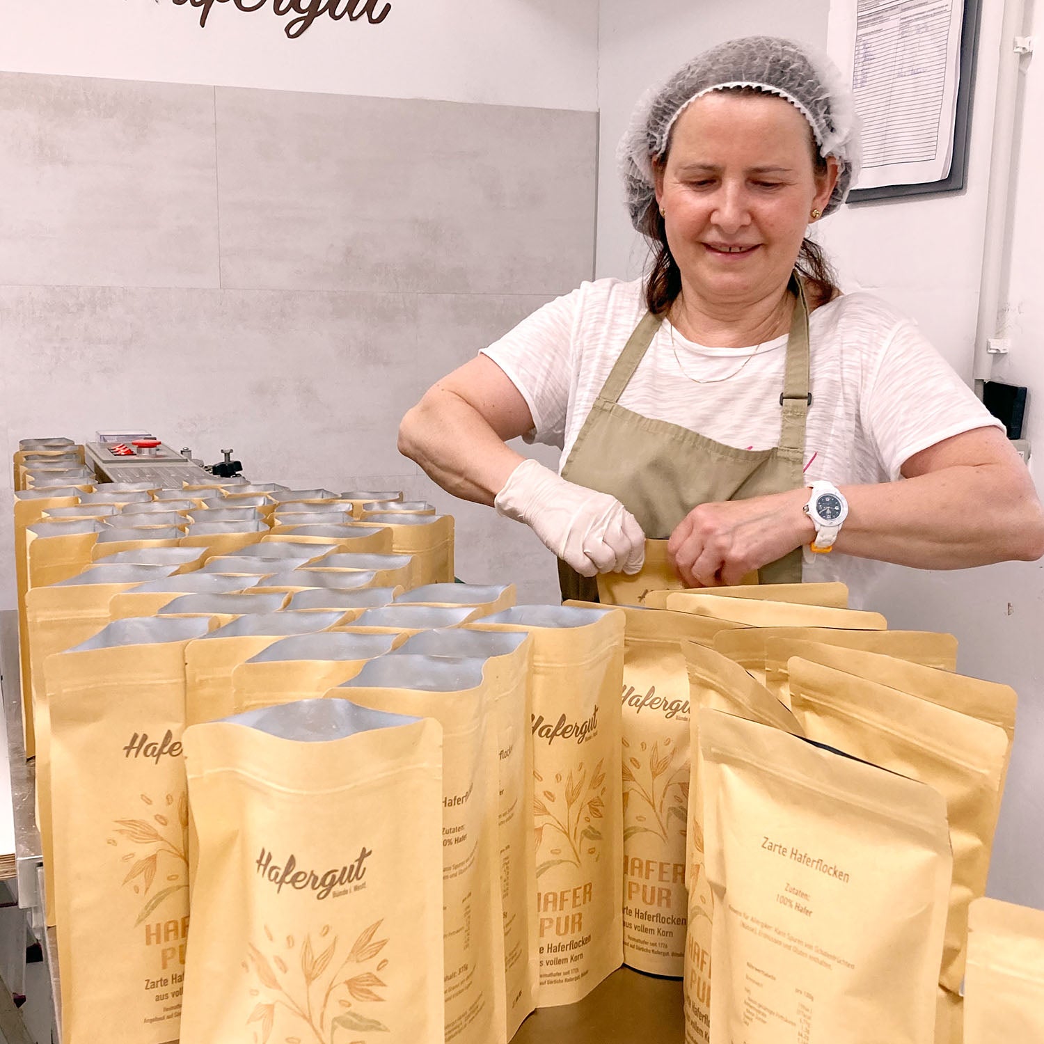 Mitarbeiterin Anna steht in der Hafergut Küche und verschließt abgefüllte Haferflocken. Sie lächelt freundlich und bestaunt dabei die Tüten, die vor ihr stehen.