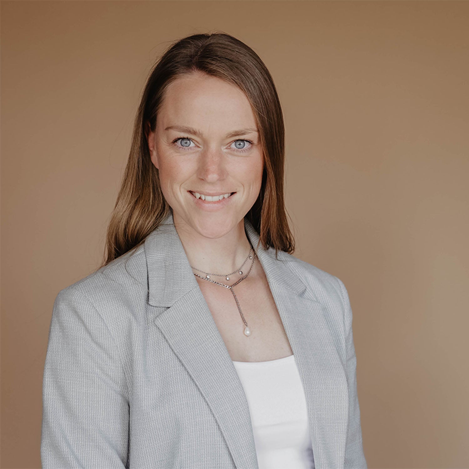 Einzelaufnahme von Jana vor einem hellbraunem Hintergrund. Sie trägt offene, dunkel blonde Hare und lächelt gerade aus in die Kamera. Sie trägt silberne Halsketten und einen Blazer in Mint.