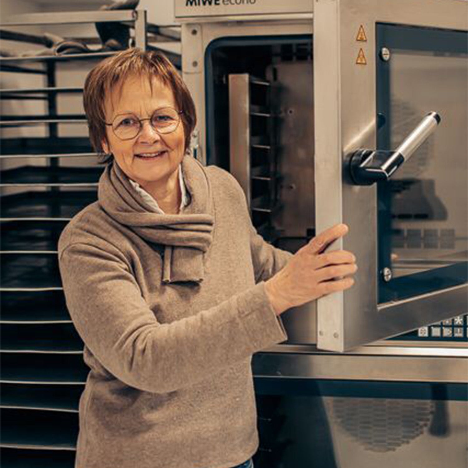 Mitarbeiterin Margitta präsentiert unseren Backofen und lächelt dabei sehr freundlich in die Kamera. Sie trägt einen samtig, braunen Pullover und dazu einen passenden Schal. Das Bild wurde von einem professionellen Fotografen aufgenommen.
